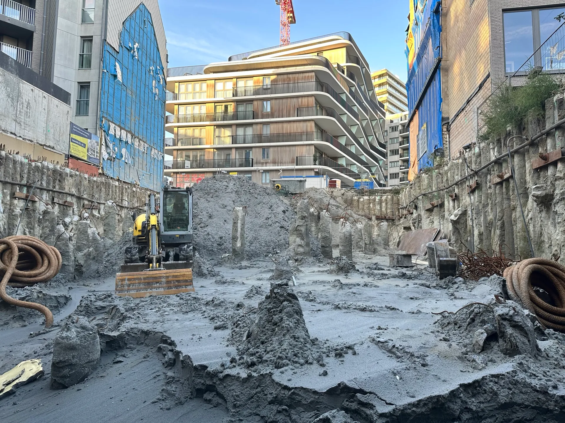 Uitgegraven bouwput met vrijstaande paalkoppen, palenwand aan weerszijden en minigraver, met appartementsgebouwen en torenkraan erachter