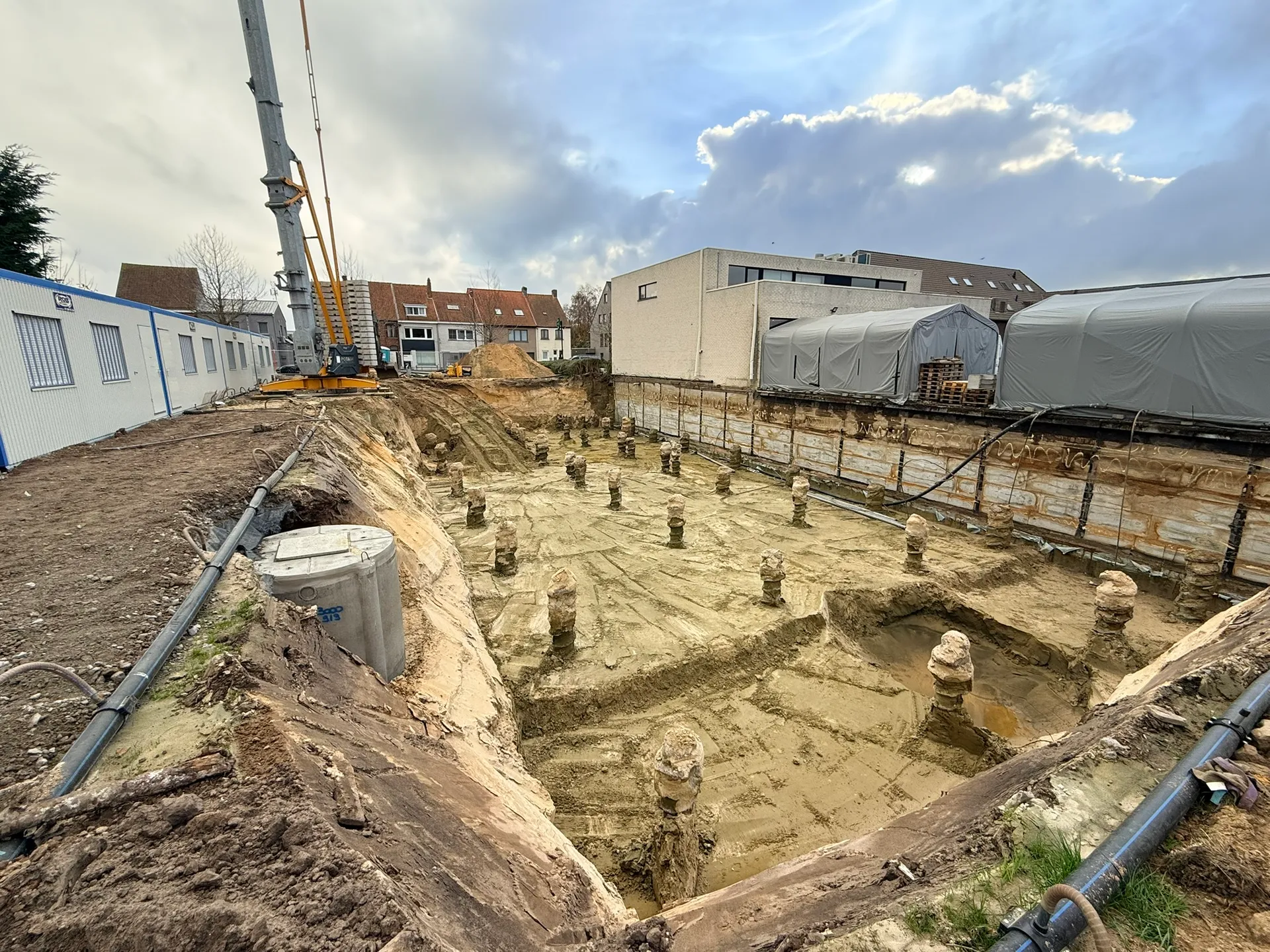 Uitgegraven bouwput met tientallen vrijgemaakte funderingspalen, een Berlinerwand langs de rechterzijde en een hoge kraan op de achtergrond