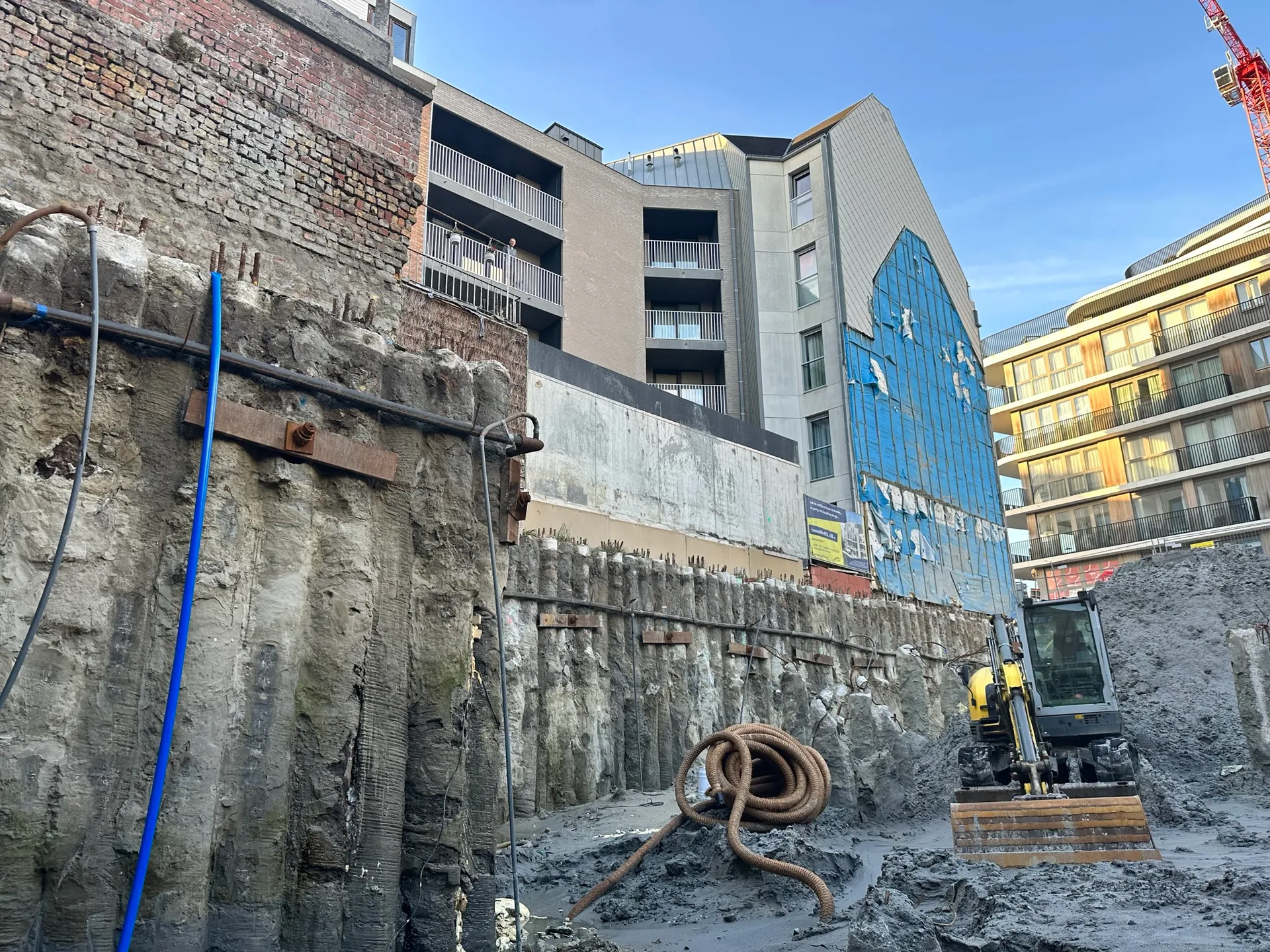Diepe bouwput met palenwand onder een bestaande bakstenen gevel, minigraver en opgerolde drainageslang tussen appartementsgebouwen