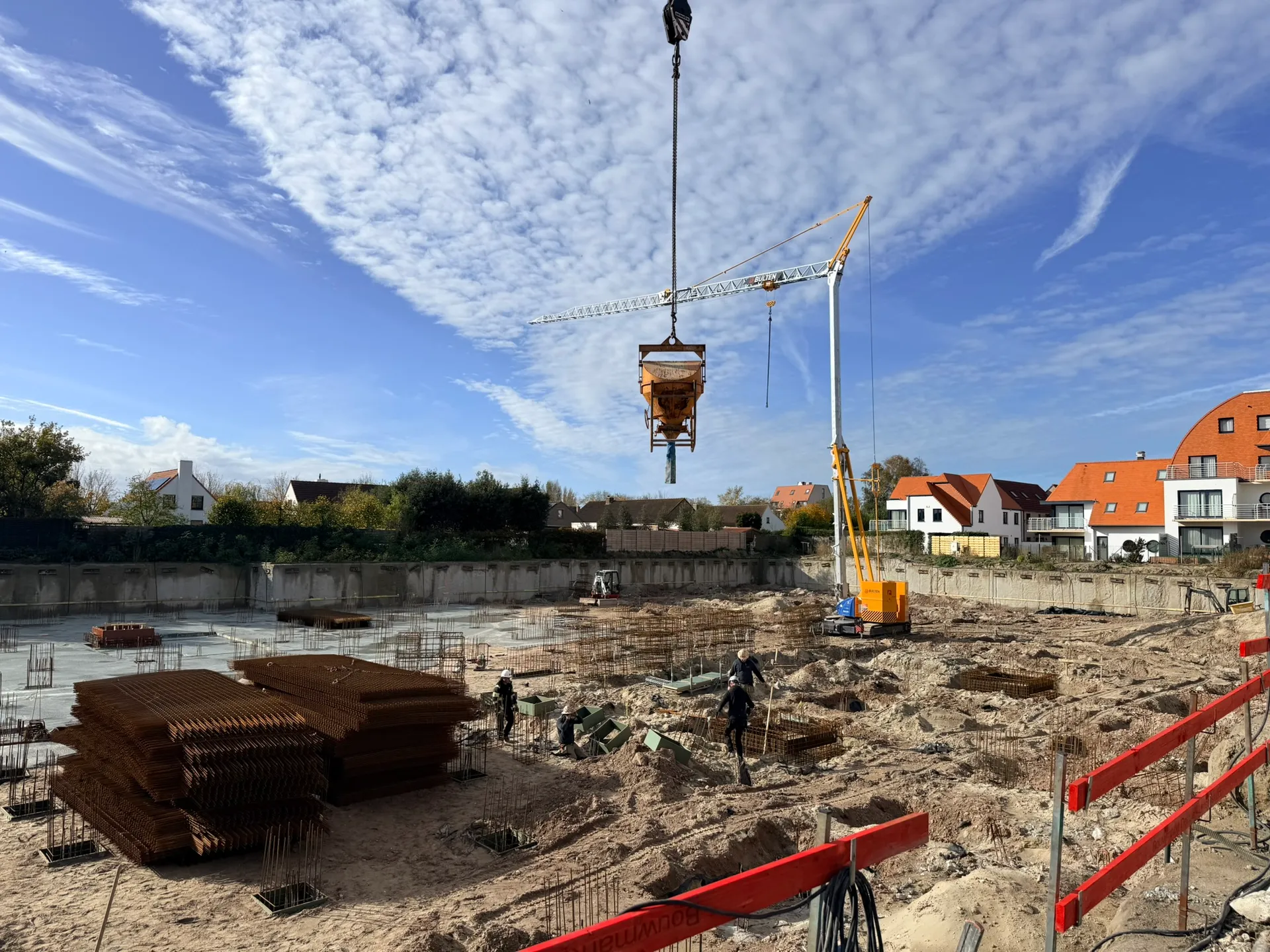 Betonkubel aan de kraan boven een bouwput met stapels wapeningsnetten en vakmannen bij wapeningskorven in het zand