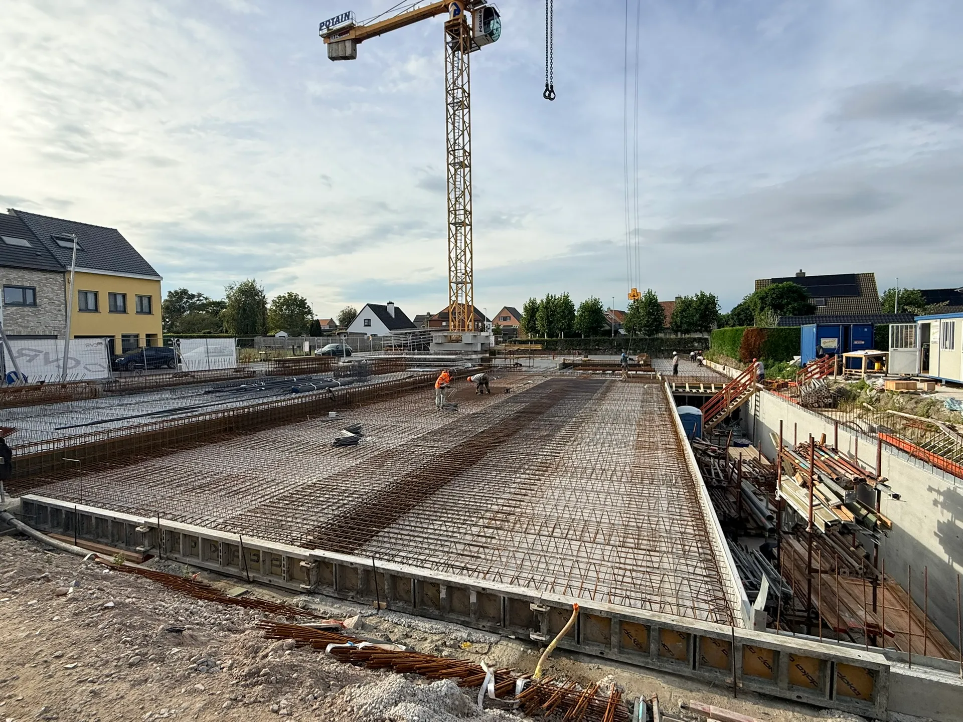 Vakmannen vlechten de wapening op het kelderdek, met randbekisting vooraan en een torenkraan boven de werf