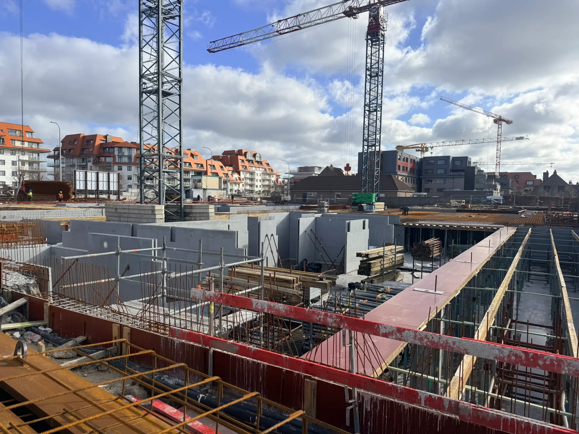 Gestorte betonnen kelderwanden naast stempels en draagbalken voor het kelderdek, met torenkranen en appartementen erachter