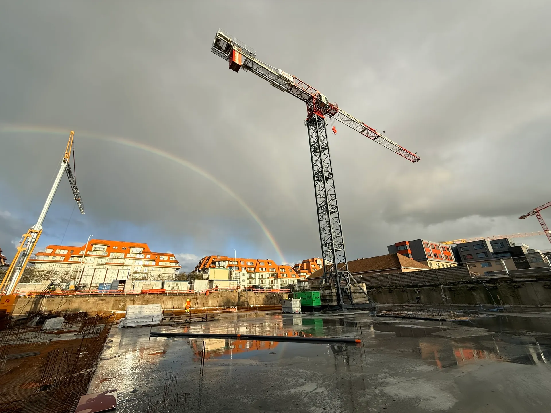 Regenboog boven een werf met torenkraan en natte, pas gestorte vloerplaat, appartementsgebouwen op de achtergrond