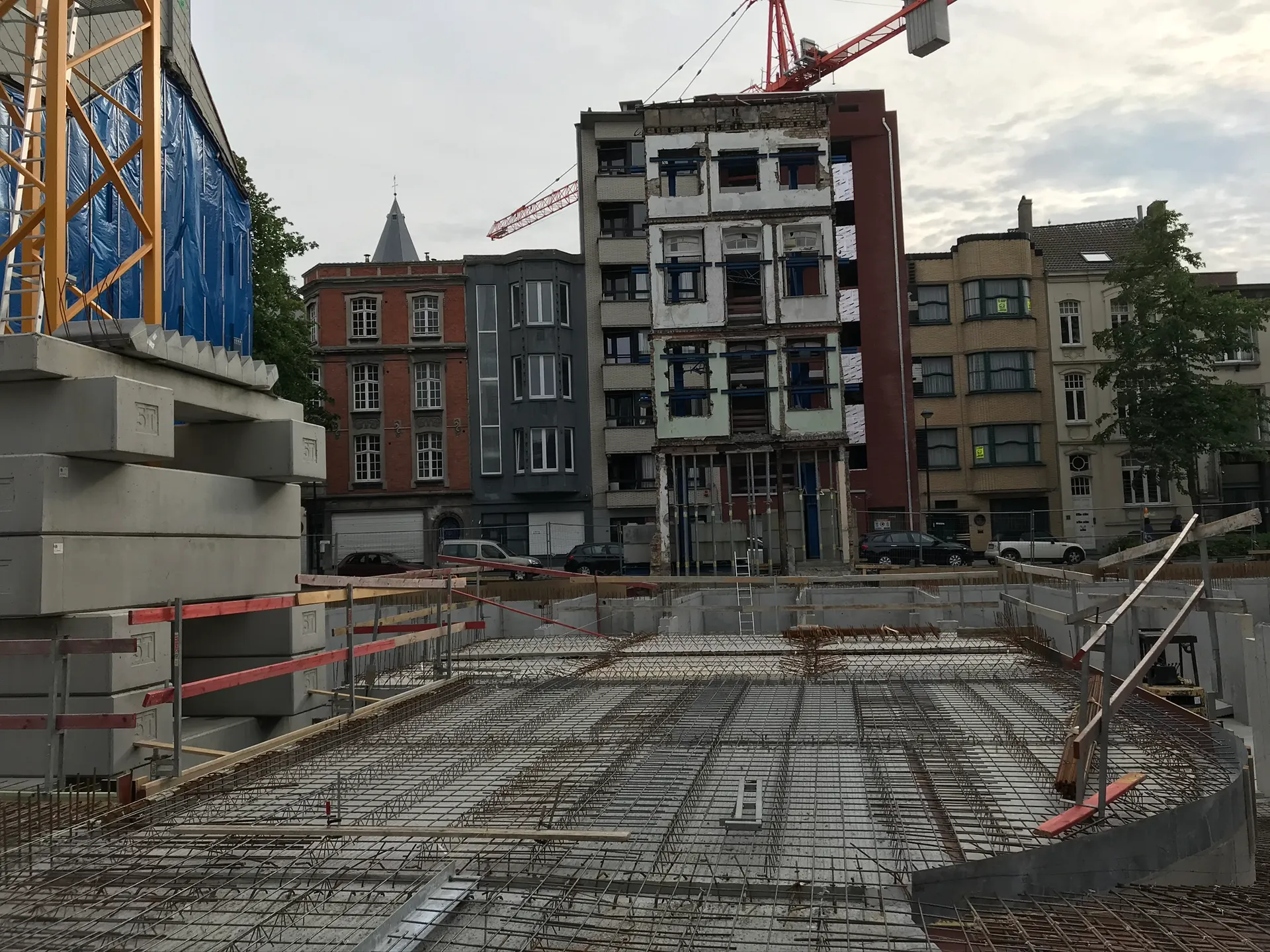 Wapening op de breedplaatvloer boven een kelder, met gestapelde prefab betonelementen en een torenkraan tussen stadsgevels