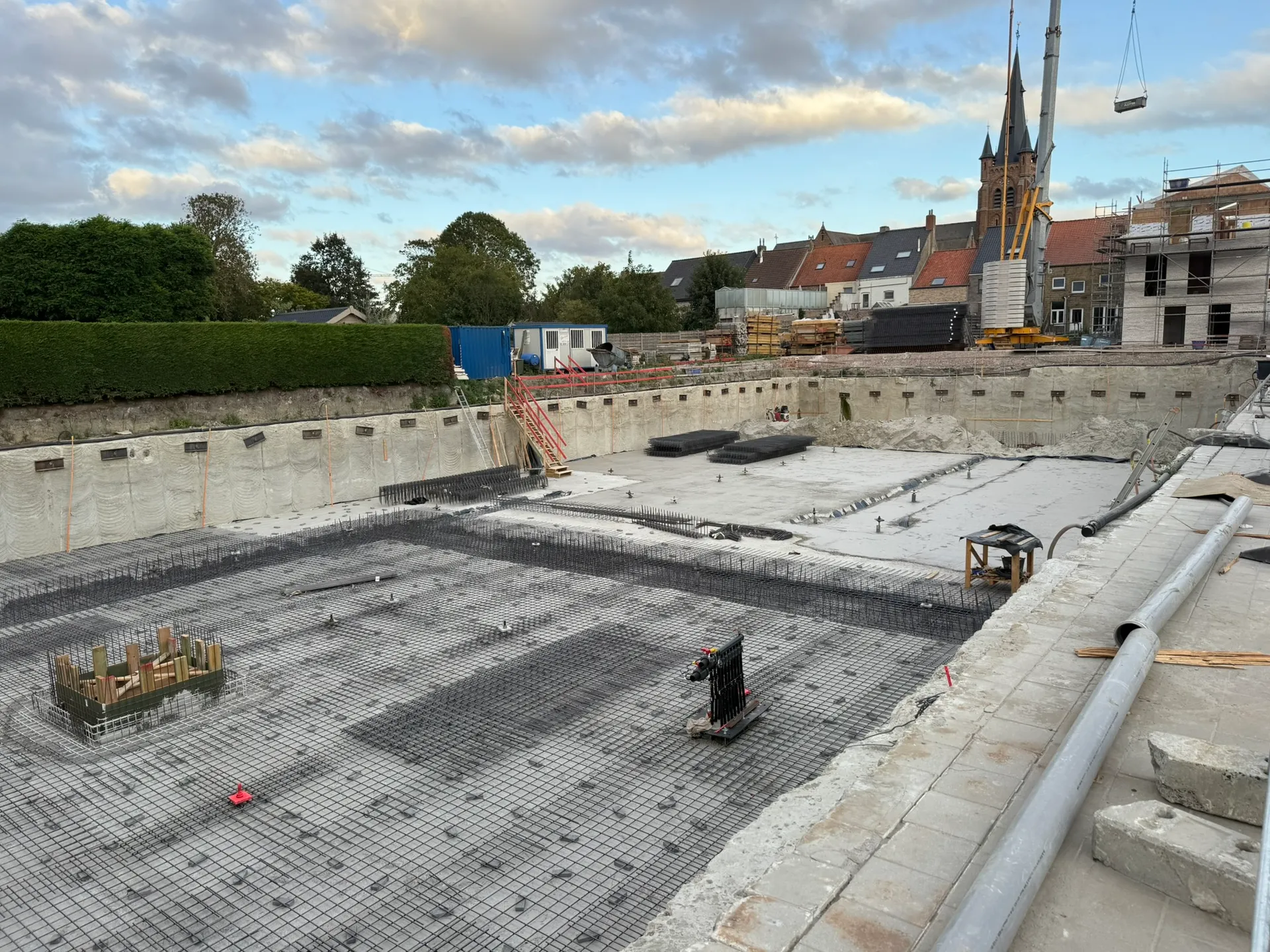 Wapeningsnetten voor de funderingsplaat in een bouwput met verankerde wanden, kerktoren en kraan op de achtergrond