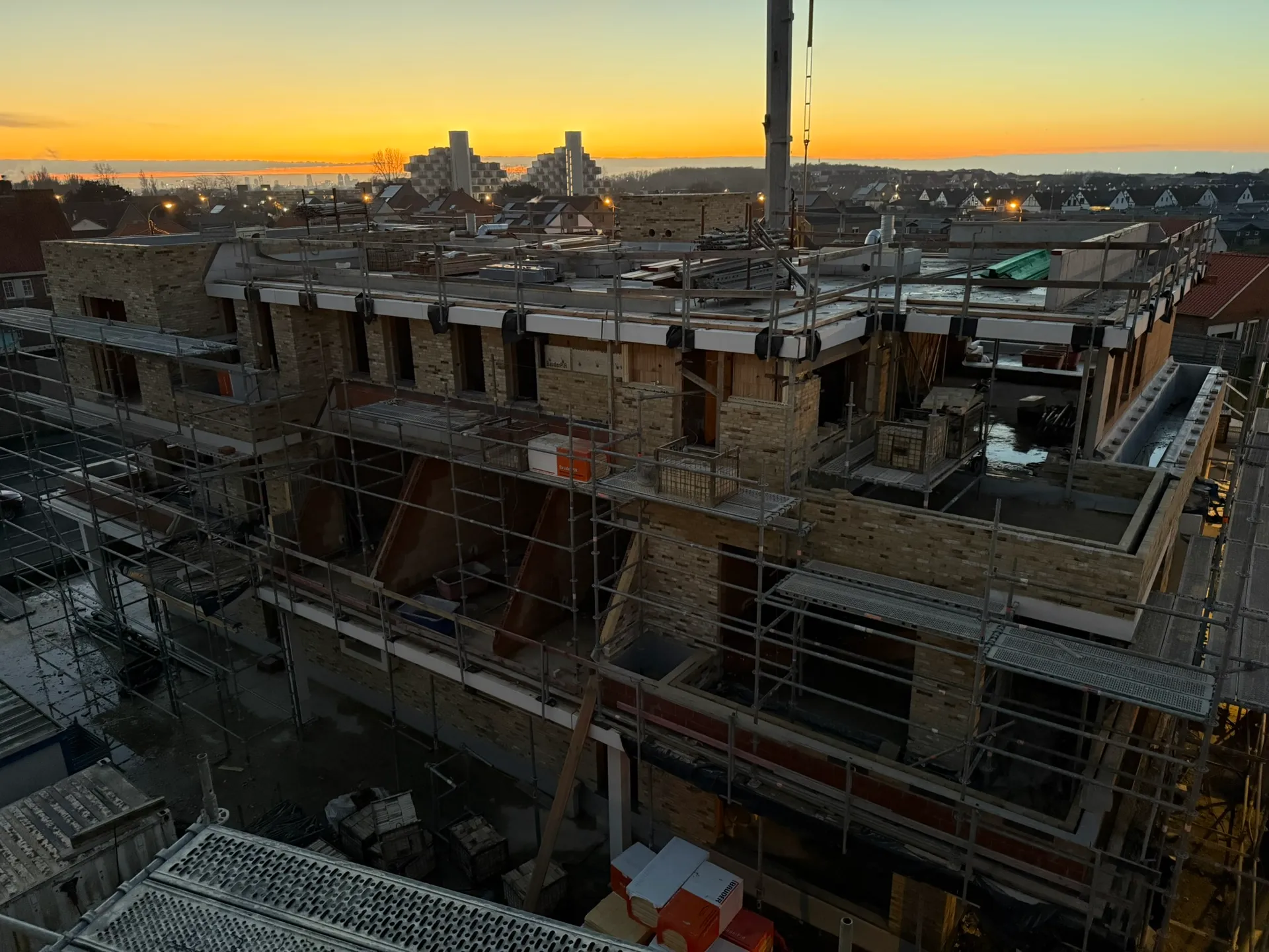 Appartementsgebouw in opbouw met steigers rond het gevelmetselwerk, van bovenaf gezien bij een oranje avondlucht