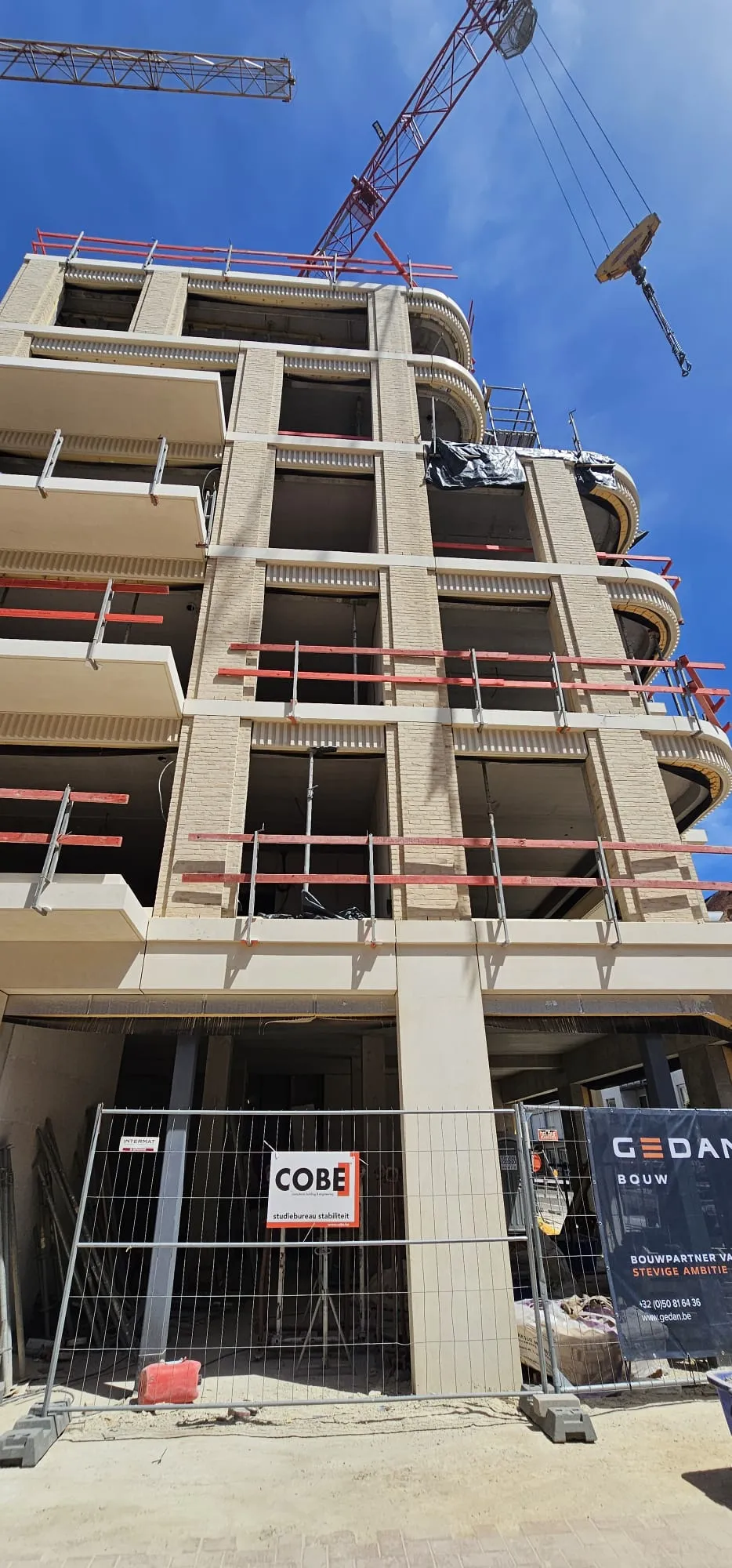 Appartementsgebouw in opbouw met afgeronde balkons en rode randbeveiliging, torenkraan erboven en GEDAN-werfdoek aan de afsluiting