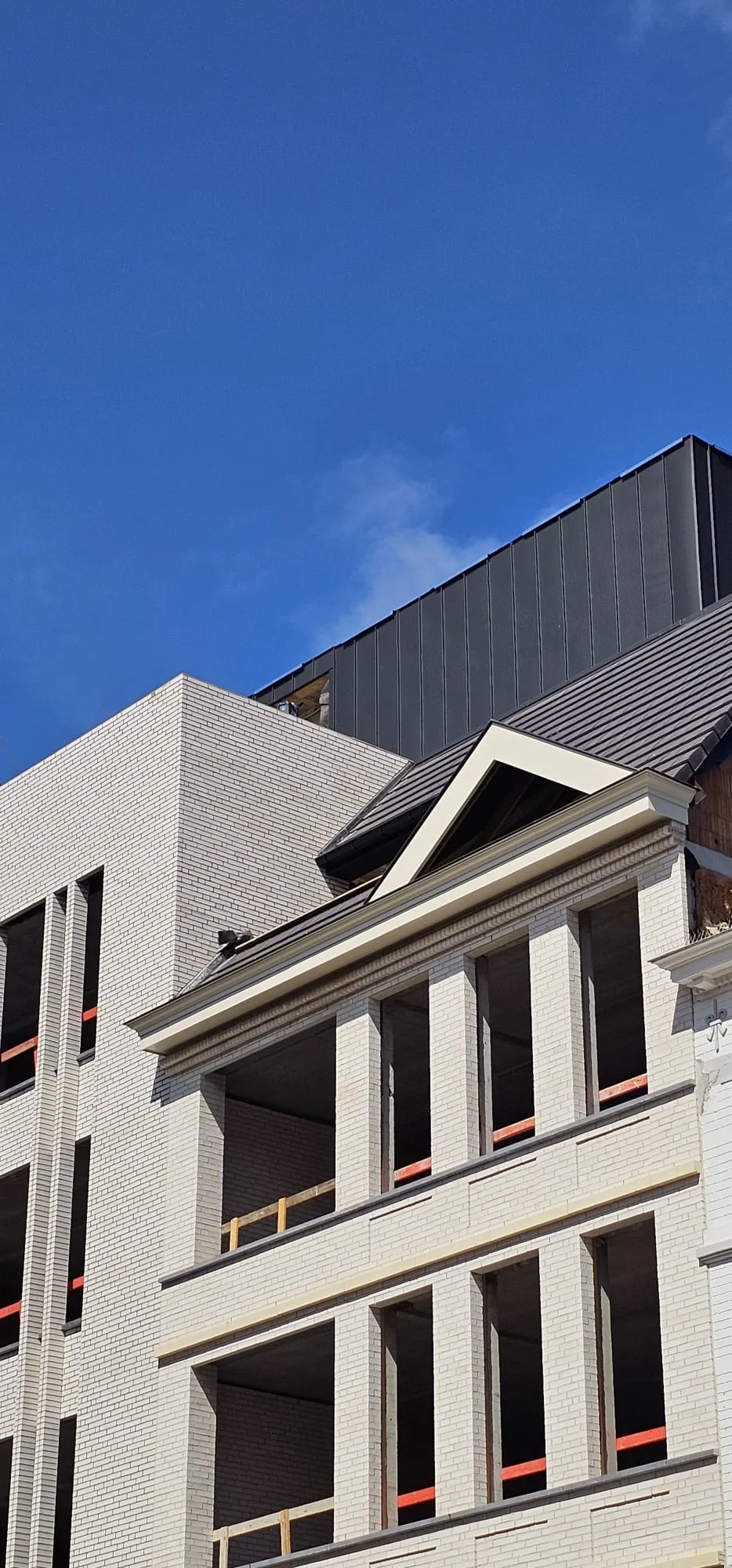 Bovenste verdiepingen van een nieuwbouw met witte gevelsteen, fronton en donker metalen dak tegen een blauwe lucht