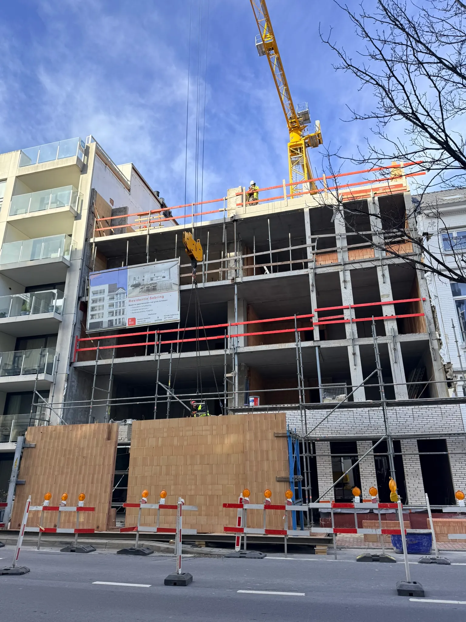 Torenkraan takelt prefab snelbouwwanden vanaf de straat naar een appartementsgebouw in ruwbouw tussen bestaande panden
