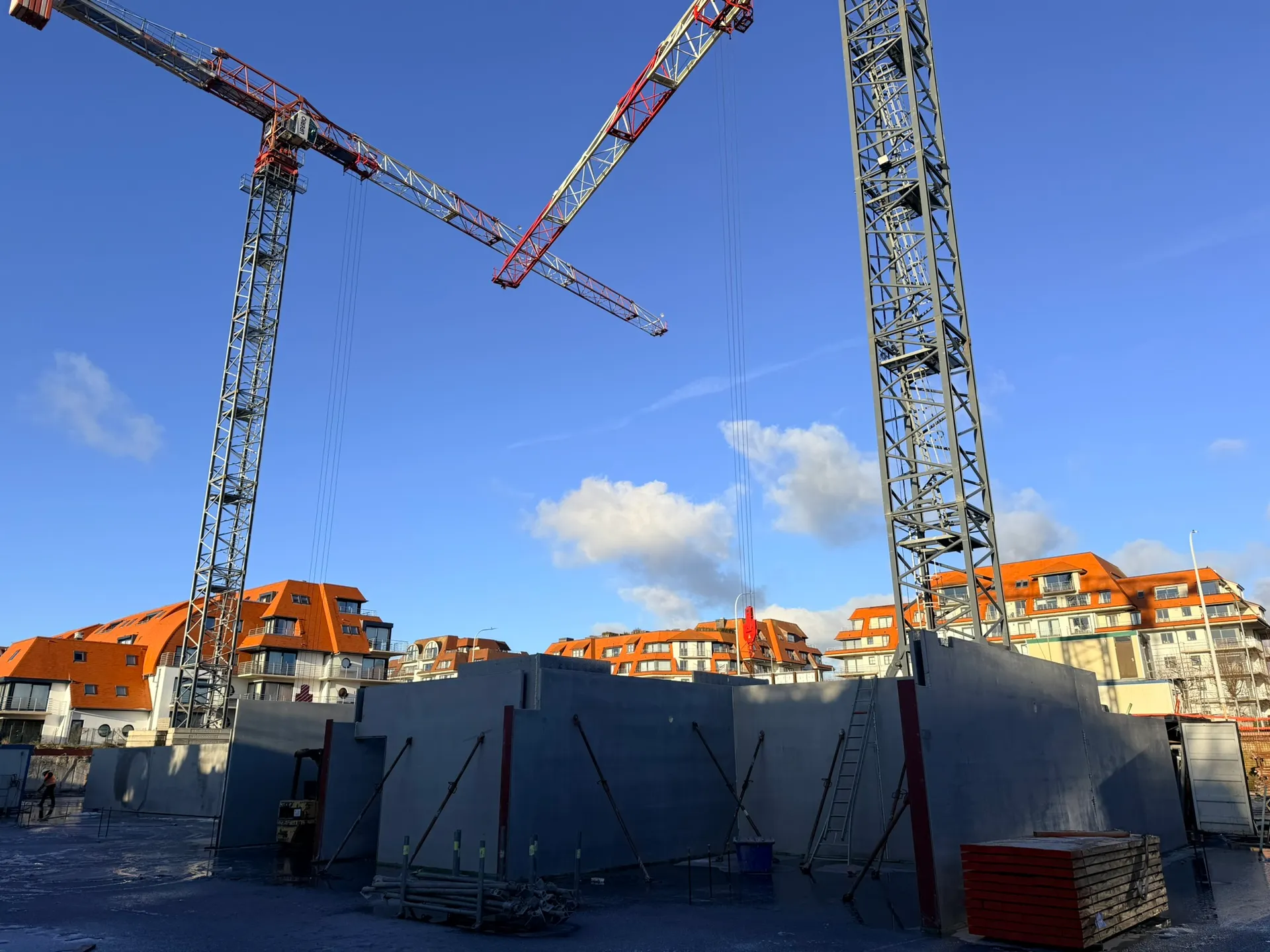 Geschoorde prefab betonwanden op de vloerplaat onder twee torenkranen, met appartementsgebouwen met oranje daken op de achtergrond