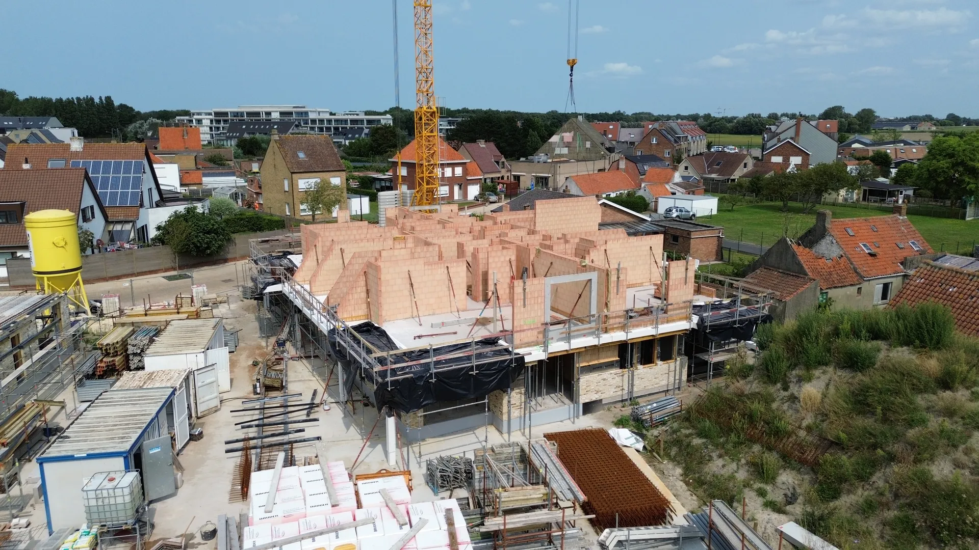 Luchtbeeld van een werf met geschoorde snelbouwmuren op de bovenste bouwlaag, gele mortelsilo, opgeslagen bouwmaterialen en torenkraan